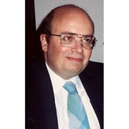 Head shot of balding male with eyeglasses, wearing black suit, white shirt, and a blue tie.