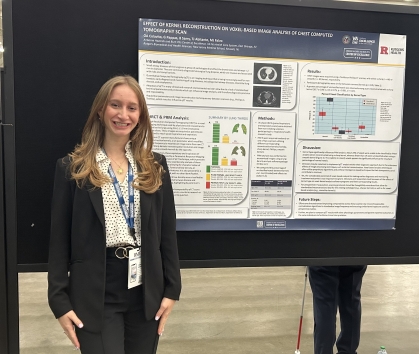 Female college student stands in front of a research poster. She has long blond hair and is wearing a black blazer with a white and black blouse.