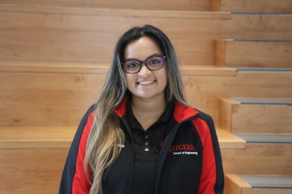 Young female college student with long brown hair parted in the middle with glasses, poses wearing a black and red zip-up jacket.