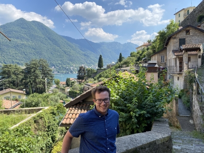 Young male wearing eyeglasses and a blue button down patterned shirt stands with a mountain, a lake, and stone and terra cotta houses in the background. 