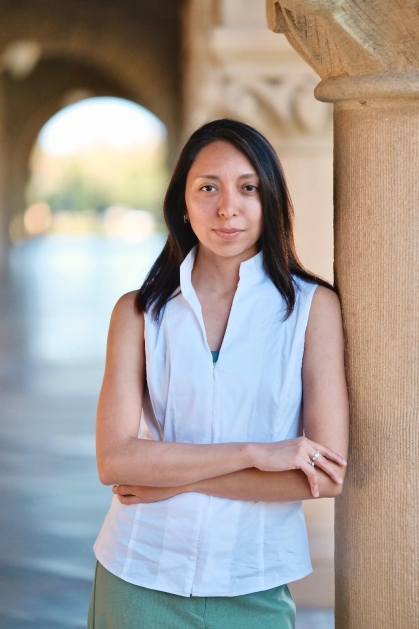 Young woman with long black hair wearing a sleeveless white blouse with her arms crossed leans against a column.