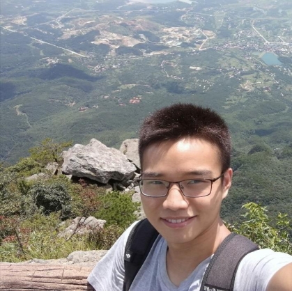 Student with short black hair wearing eyeglasses standing on a mountain with the valley in the background.