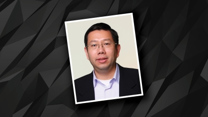 Headshot placed on black and grey patterned background of male with eyeglasses wearing a black suit jacket and white button down shirt.