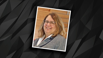 Headshot on black and grey patterned background of a female with eyeglasses and short brown hair wearing a grey suit jacket.
