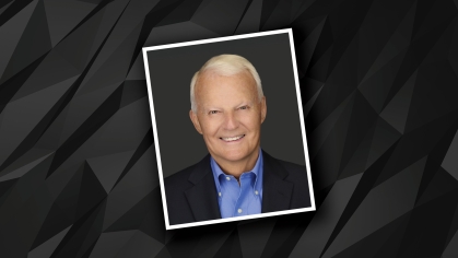Headshot of male with short white hair wearing a blue button down shirt and a black suit jacket against a black and grey patterned background