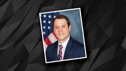 Headshot of male wearing a dark suit, white shirt, and stripped tie with an American flag in the background.