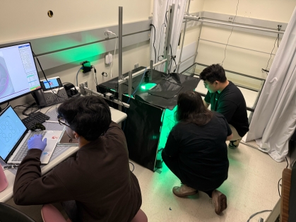 Three college students conduct lab experiment. Their backs are to the camera and one is working on a lap top while the others are illuminated under a green light.
