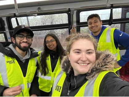 Four students pose on a bus wearing yellow safety vests.
