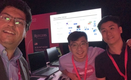 Male professor poses for a closeup with two male students with a monitor display behind them.