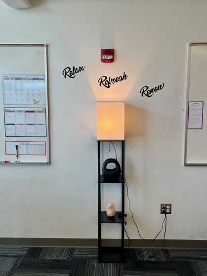 A vertical shelf with different objects and a lamp on top, with the words "Relax, Refresh, Renew" pasted on the wall above.