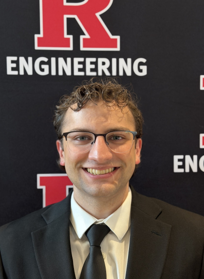 Man with short curly hair wearing a suit jacket and tie in front of an R Engineering banner backdrop