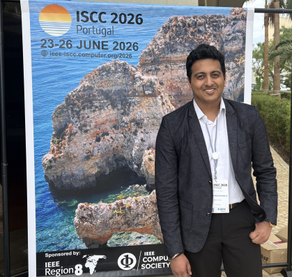 Man in dark suit with white shirt standing in front of a poster for the ISCC 2026 conference in Portugal