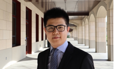 Man in dark suit, blue shirt and tie stands in corridor with archways behind him.