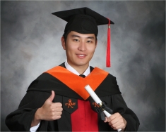 Male student of Chinese dissent wearing a graduation cap and gown holding a diploma and giving a thumbs up sign.