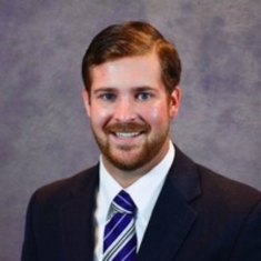 Head shot of male with brown hair, a moustache and a beard, wearing a black suit jacket, a white shirt, and a blue patterned tie.