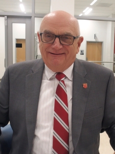 Headshot of bald male with eyeglasses wearing a grey suit, shite button down shirt, and red patterned tie.