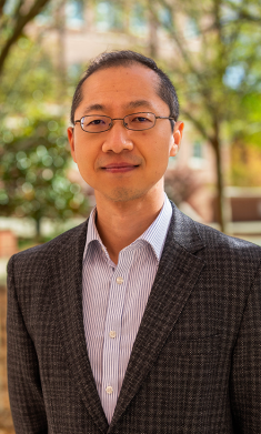 Head shot of male with eyeglasses wearing a dark brown patterned suit with a button down shirt.