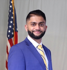 Headshot of male with short black hair and a beard wearing a blue suit, white button down shirt, and yellow patterned tie with a U.S. flag in the background.