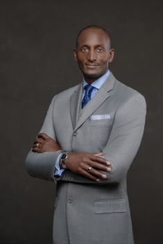 Black male wearing a light gray suit with a light blue shirt and dark blue tie stands against a dark background with arms folded.