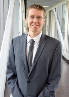 Head shot of male with eyeglasses wearing a grey suit and tie with a white button down shirt.