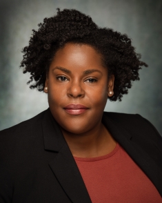 Headshot of Black woman with short dark hair and pearl earrrings, wearing a black suit jacket with a brown sweater.