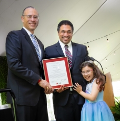 Man on left wearing a dark suit presents an award to a male faculty member alongside his young daughter wearing a light blue dress..