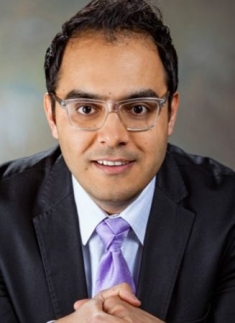 Head shot of male with eyeglasses, wearing a black suit, purple shirt, and purple tie.
