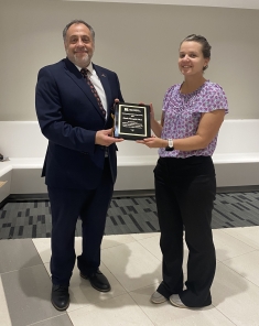 A man and a woman pose holding a plaque. The man is wearing a suit and the woman is wearing black pants and a purple blouse.