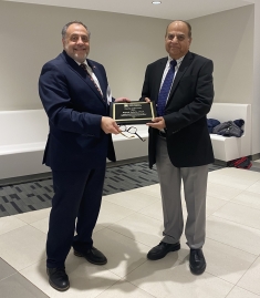 Two men wearing suits stand together holding a plaque.