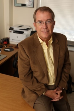 Headshot of male with short hair, wearing eyeglasses, a brown suit jacket with a yellow button down shirt.