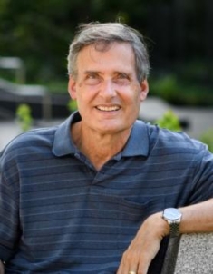 Headshot of male with short grey hair, wearing a blue polo shirt and a wrist watch.