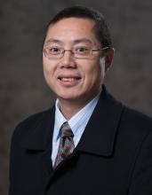 Headshot of Asian male with eyeglasses, wearing a black suit with a light blue shirt, and patterned tie.