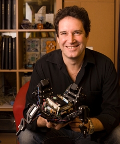 Headshot of male with short, curly brown hair, holding a small robot.