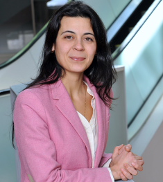 Woman with long brown hair, wearing a pink jacket with a white v-neck shirt.