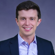 Headshot of male with short black hair wearing a black suit jacket and a blue button down shirt.